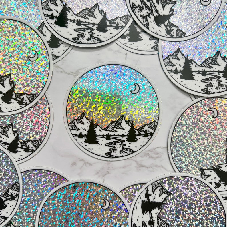 Puede incluir: Pegatinas circulares con un fondo hologr&aacute;fico y un dise&ntilde;o de paisaje de monta&ntilde;as y bosques en blanco y negro. Cada pegatina presenta una luna creciente y una ilustraci&oacute;n detallada de &aacute;rboles, monta&ntilde;as y un r&iacute;o.