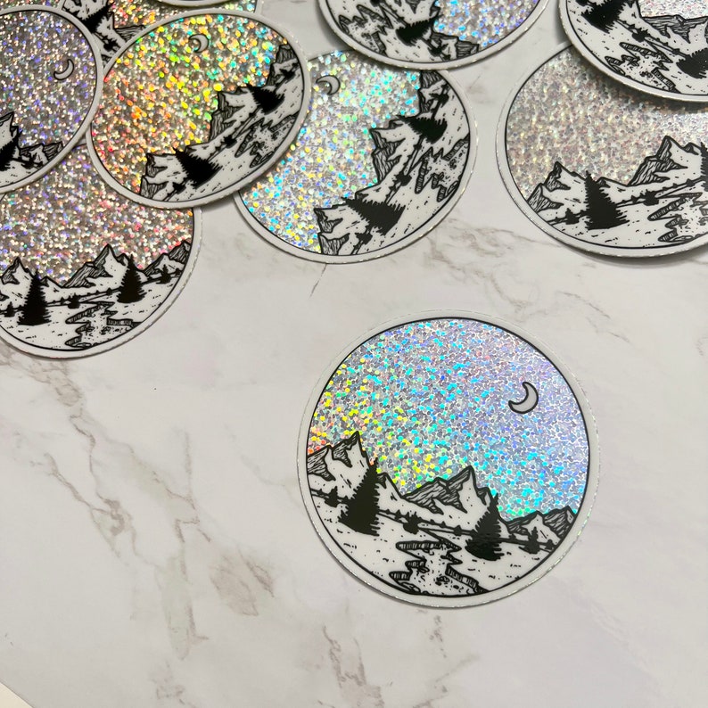 Puede incluir: Pegatinas hologr&aacute;ficas circulares con un paisaje monta&ntilde;oso en blanco y negro y una luna creciente. El dise&ntilde;o incluye &aacute;rboles, un r&iacute;o y monta&ntilde;as. Las pegatinas tienen un efecto brillante e iridiscente.