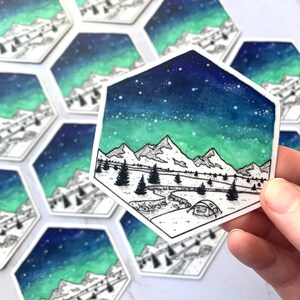 May include: A hexagonal sticker with a watercolor-style illustration of a mountain range under a starry sky with a green and blue aurora borealis. A small tent is in the foreground.