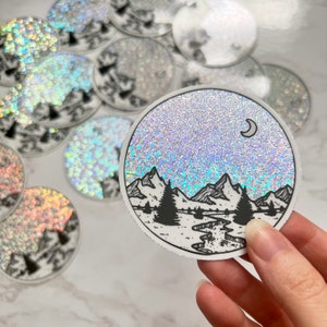 Puede incluir: Pegatina redonda con un dise&ntilde;o de paisaje de monta&ntilde;as y bosques. La pegatina presenta una ilustraci&oacute;n en blanco y negro de monta&ntilde;as, &aacute;rboles y un r&iacute;o, sobre un fondo brillante y brillante con una luna creciente.