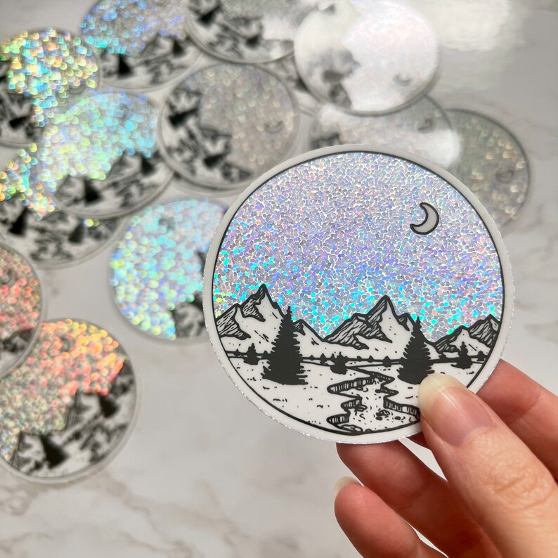 Puede incluir: Pegatina redonda con un dise&ntilde;o de paisaje de monta&ntilde;as y bosques. La pegatina presenta una ilustraci&oacute;n en blanco y negro de monta&ntilde;as, &aacute;rboles y un r&iacute;o, sobre un fondo brillante y brillante con una luna creciente.