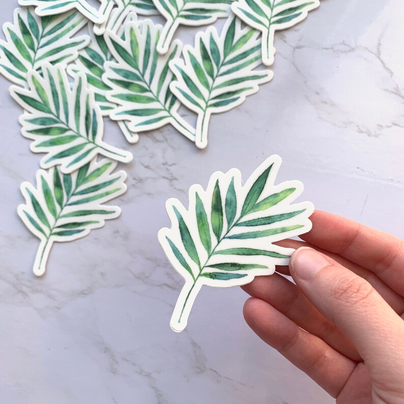 May include: A set of green watercolour palm leaf stickers. The stickers are on a white marble surface. One sticker is being held by a hand.
