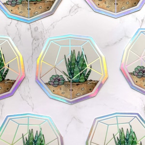 Könnte beinhalten: Ein Set mit sechs schimmernden Aufklebern, die ein geometrisches Terrarium-Design mit einer Sukkulentenpflanze im Inneren zeigen. Die Aufkleber haben eine holografische Oberfläche und sind perfekt, um jeder Oberfläche einen Hauch von Verspieltheit zu verleihen.