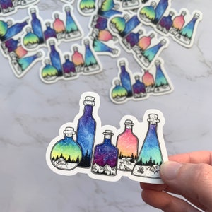 May include: Five glass bottles with cork stoppers, each containing a different watercolor scene of a starry night sky, mountains, and a forest. The bottles are arranged in a row on a white surface.