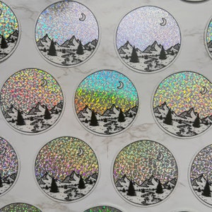 Puede incluir: Pegatinas hologr&aacute;ficas circulares con un paisaje de monta&ntilde;a en blanco y negro y una luna creciente. El dise&ntilde;o incluye &aacute;rboles, un r&iacute;o y monta&ntilde;as, con un efecto brillante y brillante. Las pegatinas est&aacute;n dispuestas sobre una superficie blanca.