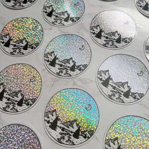 Puede incluir: Pegatinas circulares con un dise&ntilde;o de monta&ntilde;a y luna. Las pegatinas presentan una ilustraci&oacute;n en blanco y negro de una cordillera, &aacute;rboles y una luna creciente. El fondo tiene un efecto hologr&aacute;fico y brillante.