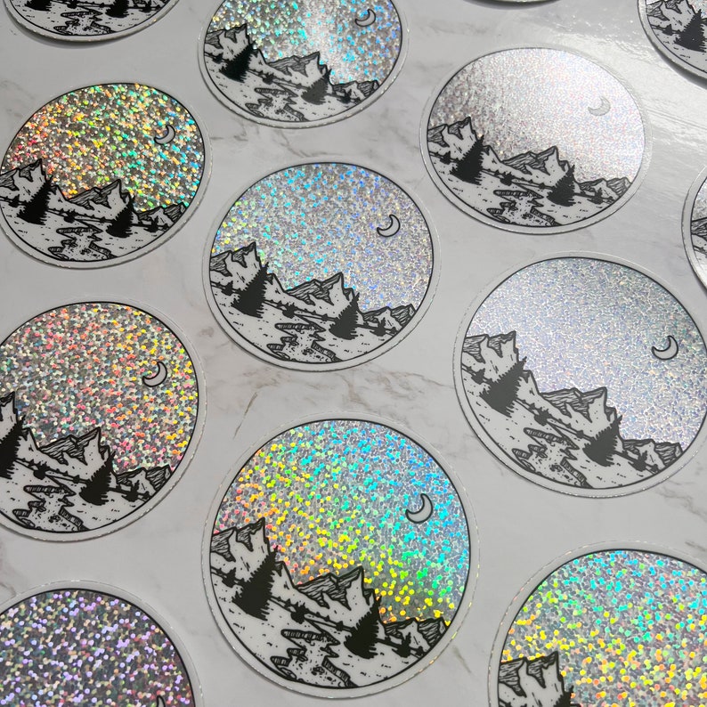 Puede incluir: Pegatinas circulares con un dise&ntilde;o de monta&ntilde;a y luna. Las pegatinas presentan una ilustraci&oacute;n en blanco y negro de una cordillera, &aacute;rboles y una luna creciente. El fondo tiene un efecto hologr&aacute;fico y brillante.