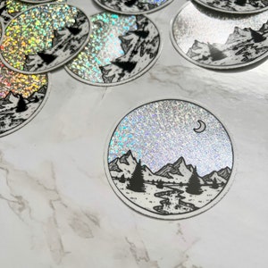 Puede incluir: Pegatinas hologr&aacute;ficas circulares con un dise&ntilde;o de paisaje de monta&ntilde;a en blanco y negro. El dise&ntilde;o incluye monta&ntilde;as, &aacute;rboles, un r&iacute;o y una luna creciente. Las pegatinas tienen un efecto brillante e iridiscente.