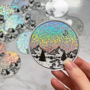 Puede incluir: Una pegatina redonda con un fondo hologr&aacute;fico y un dise&ntilde;o de paisaje de monta&ntilde;a en blanco y negro. El dise&ntilde;o incluye monta&ntilde;as, &aacute;rboles, un r&iacute;o y una luna creciente. Se ven otras pegatinas similares.