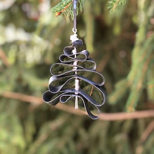 May include: A black metal Christmas tree ornament made from recycled inner tubes. The ornament is hanging from a branch of a pine tree.