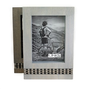 May include: A gray wooden picture frame with a silver chain detail on the bottom. The frame has a black and white photo of a person on a bicycle in a mountainous landscape. The photo has the text "LINKS" on the bottom.