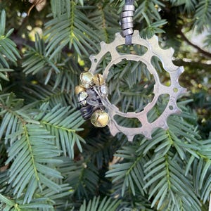 May include: A silver bicycle chainring with a black leather cord and a cluster of gold and silver bells hanging from it. The chainring is hanging from a green evergreen tree branch.