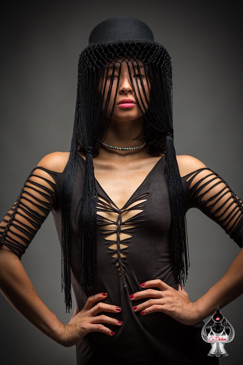 Puede incluir: Una mujer con un top negro con un dise&ntilde;o de corte y un sombrero negro con flecos. El sombrero le cubre la cara.