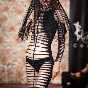 Puede incluir: Una modelo lleva un vestido negro, transparente, con recortes, mangas largas y un sombrero negro con velo de flecos. El vestido tiene un corpi&ntilde;o ajustado y una falda fluida. La modelo camina por una pasarela con un fondo floral.