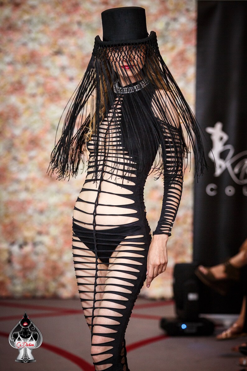 Puede incluir: Una modelo lleva un vestido negro, transparente, con recortes, mangas largas y un sombrero negro con velo de flecos. El vestido tiene un corpi&ntilde;o ajustado y una falda fluida. La modelo camina por una pasarela con un fondo floral.
