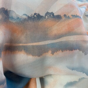 Puede incluir: Primer plano de una tela fluida con un dise&ntilde;o de acuarela. La tela presenta una escena de paisaje con tonos de azul, naranja y gris. El dise&ntilde;o se asemeja a una puesta de sol sobre una masa de agua.