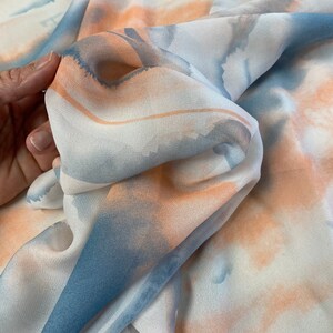 Puede incluir: Primer plano de una tela fluida con un dise&ntilde;o de acuarela. La tela presenta suaves tonos de azul, melocot&oacute;n y blanco, creando un efecto et&eacute;reo. El material parece ligero y delicado, con pliegues y texturas sutiles.