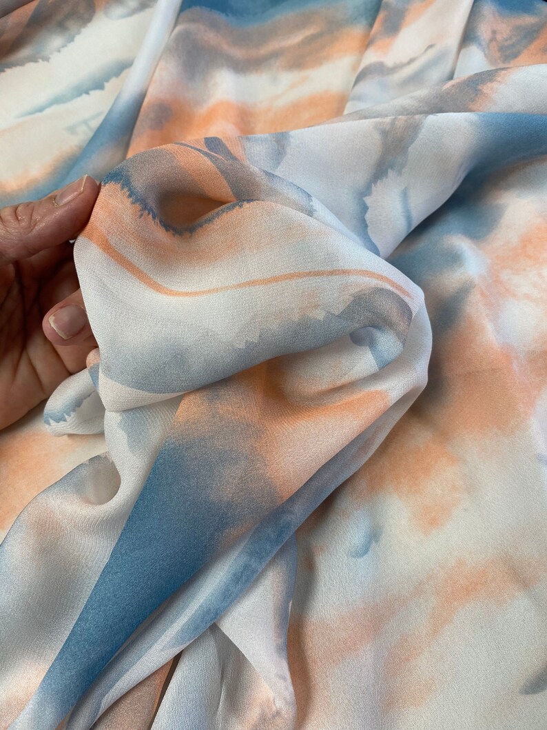 Puede incluir: Primer plano de una tela fluida con un dise&ntilde;o de acuarela. La tela presenta suaves tonos de azul, melocot&oacute;n y blanco, creando un efecto et&eacute;reo. El material parece ligero y delicado, con pliegues y texturas sutiles.