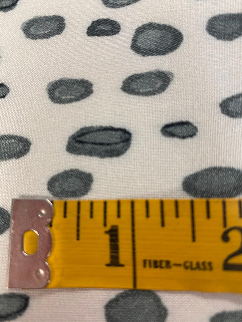 May include: A close-up of a yellow measuring tape with black markings. The tape reads "1" and "2" and has the words "Fibre-Glass" printed on it. The tape is lying on a white fabric with a black and white polka dot pattern.