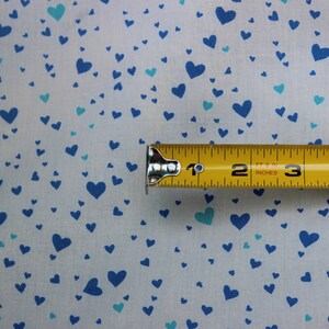 May include: A white fabric with a blue and turquoise heart pattern. A yellow measuring tape is placed on top of the fabric, showing measurements in inches and feet.
