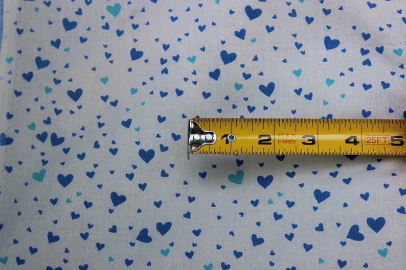 May include: A white fabric with a blue and turquoise heart pattern. A yellow measuring tape is placed on top of the fabric, showing measurements in inches and feet.