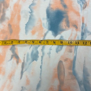 Puede incluir: Tela blanca con un dise&ntilde;o abstracto de acuarela en tonos melocot&oacute;n y azul. Una cinta m&eacute;trica amarilla est&aacute; colocada sobre la tela, mostrando medidas de hasta 33 cm.