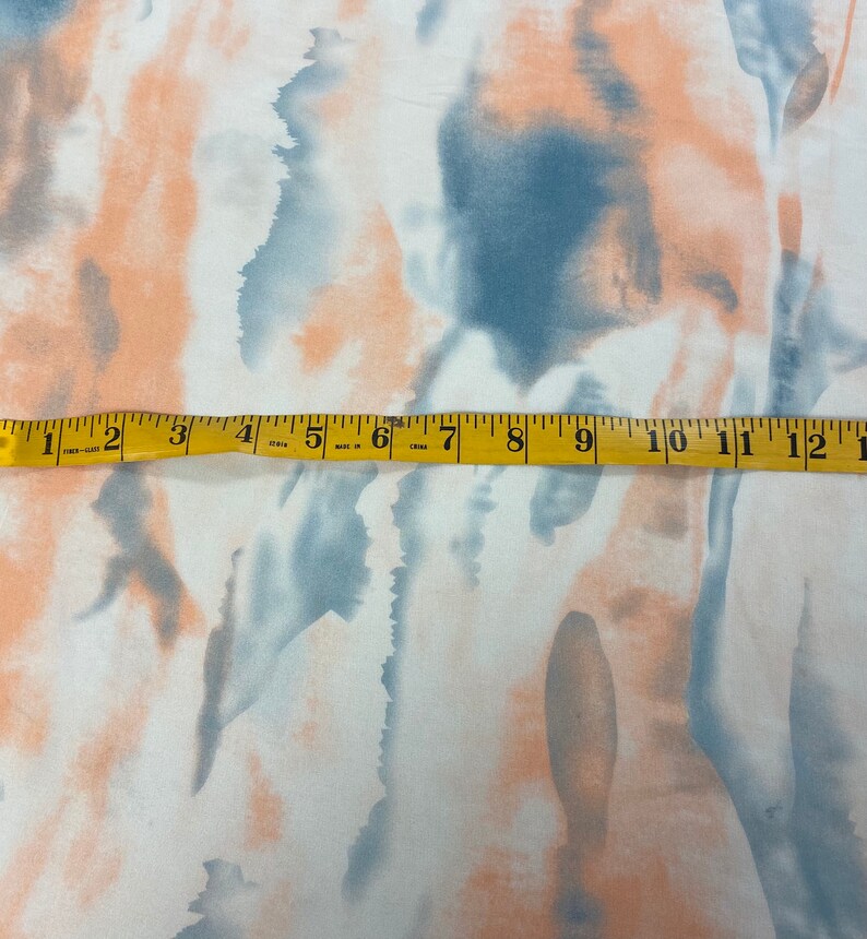Puede incluir: Tela blanca con un dise&ntilde;o abstracto de acuarela en tonos melocot&oacute;n y azul. Una cinta m&eacute;trica amarilla est&aacute; colocada sobre la tela, mostrando medidas de hasta 33 cm.