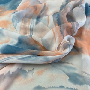 Puede incluir: Una tela fluida con un dise&ntilde;o tipo acuarela. El patr&oacute;n presenta suaves tonos mezclados de azul, melocot&oacute;n y blanco, creando un efecto et&eacute;reo y de ensue&ntilde;o. La tela est&aacute; drapeada, mostrando su textura y la interacci&oacute;n de los colores.