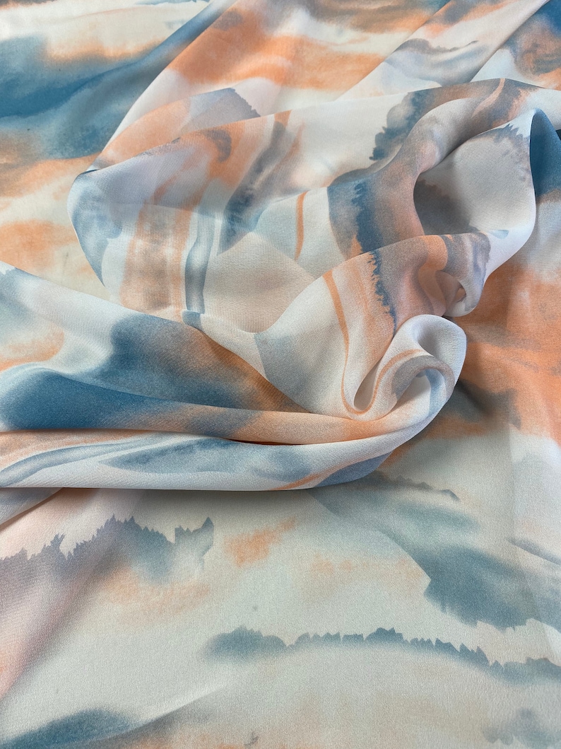 Puede incluir: Una tela fluida con un dise&ntilde;o tipo acuarela. El patr&oacute;n presenta suaves tonos mezclados de azul, melocot&oacute;n y blanco, creando un efecto et&eacute;reo y de ensue&ntilde;o. La tela est&aacute; drapeada, mostrando su textura y la interacci&oacute;n de los colores.