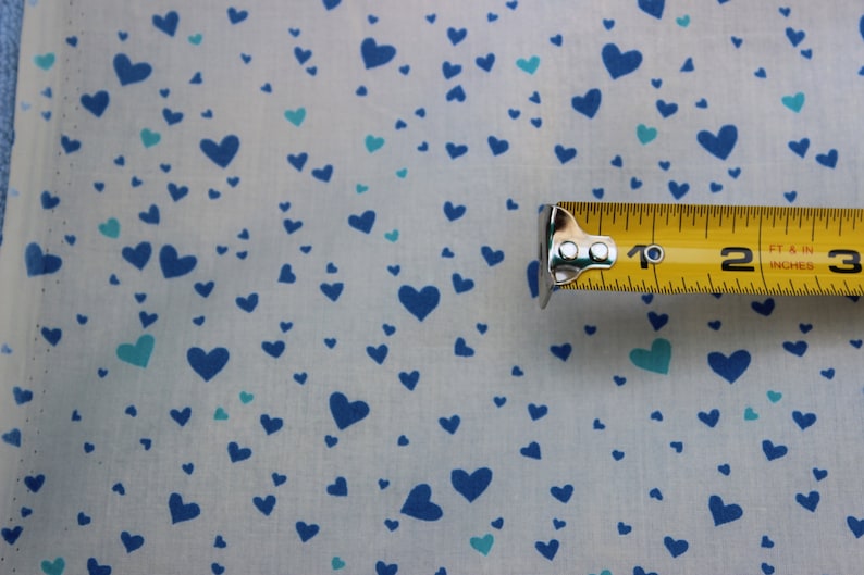 May include: White fabric with a blue and teal heart pattern. A yellow measuring tape with the numbers "1", "2", and "3" is visible.