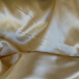 Puede incluir: Primer plano de una tela suave y sedosa de color amarillo crema p&aacute;lido. La tela est&aacute; drapeada y doblada, creando l&iacute;neas suaves y fluidas y una sensaci&oacute;n de lujo.