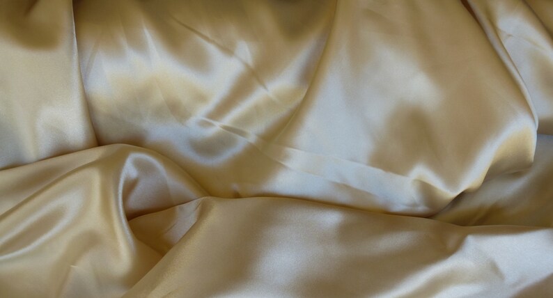 Puede incluir: Primer plano de una tela suave y sedosa de color amarillo crema p&aacute;lido. La tela est&aacute; drapeada y doblada, creando l&iacute;neas suaves y fluidas y una sensaci&oacute;n de lujo.