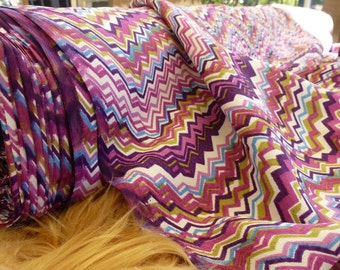 May include: A roll of fabric with a vibrant zigzag pattern in shades of purple, pink, blue, green, and white. The fabric appears to be lightweight and suitable for clothing or home decor projects. The fabric is on a roll.