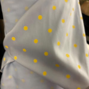 Puede incluir: Una tela de color azul claro con un patrón repetitivo de pequeños puntos amarillos sólidos. El material está drapeado, creando pliegues y sombras suaves que resaltan la textura lisa y la distribución constante de los puntos amarillos circulares.