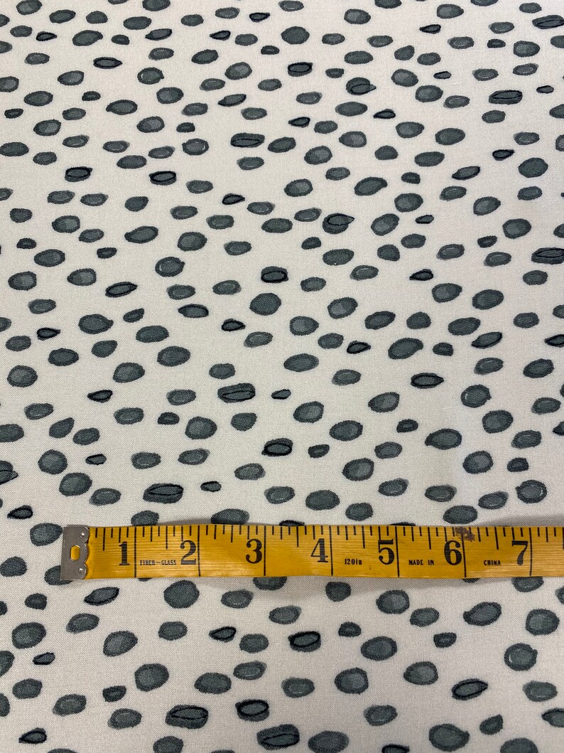May include: White fabric with a repeating pattern of small, dark grey oval shapes. A yellow measuring tape with black markings is placed across the fabric. The tape reads "1 2 3 4 120in 5 MADE IN 6 CHINA 7".
