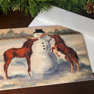 May include: A Christmas card featuring a snowman with a black top hat and a carrot nose. Two brown foals are interacting with the snowman in a snowy winter scene.