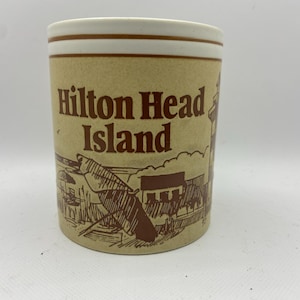 May include: Tan ceramic mug with the words "Hilton Head Island" in brown lettering. The mug features a brown illustration of a beach scene with a lighthouse and buildings. The rim has a white and brown band.