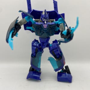 May include: A blue and teal action figure of a robot with articulated joints. The figure has a detailed design with a transparent chest and teal accents on the arms and legs. The robot is posed with its arms outstretched.
