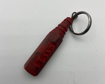 May include: A red, vintage Esso keychain. The keychain is shaped like a bottle and has the word "ESSO" printed on it. The keychain is attached to a silver metal ring.