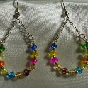 Puede incluir: Un par de pendientes de cuentas de cristal de colores arcoíris con pendientes de cadena de plata. Las cuentas están en orden de arcoíris, comenzando con verde, amarillo, naranja, rojo, rosa, azul y terminando con verde.