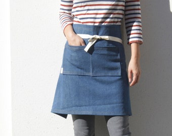 May include: A denim apron with a white tie, featuring a large front pocket. The model is wearing a striped long-sleeve shirt with red, white, and blue stripes, gray pants, and black shoes. The apron is a medium blue color.