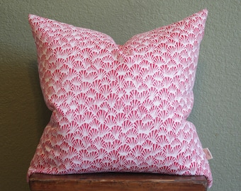 May include: A square decorative pillow with a red and white fan-shaped pattern. The pillow is resting on a wooden surface. The pillow's fabric has a repeating design of stylized scallop shells.