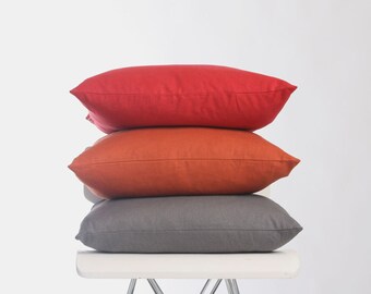 May include: Three rectangular throw pillows stacked on top of each other. The top pillow is red, the middle pillow is orange, and the bottom pillow is gray. The pillows are on a white chair.