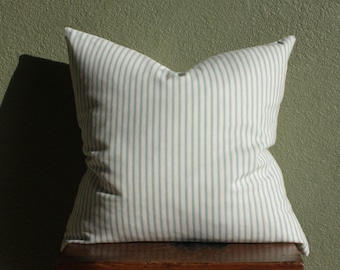May include: A square pillow with a cream-colored background and vertical stripes in a light blue-green color. The pillow is placed on a wooden stool against a textured green wall.