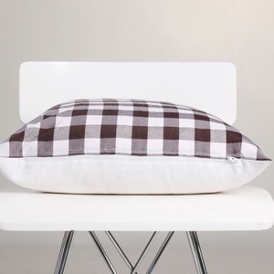 May include: A rectangular pillow with a brown and white checkered pattern on top, resting on a white pillow base. The pillow is placed on a white chair with metal legs, against a neutral background.