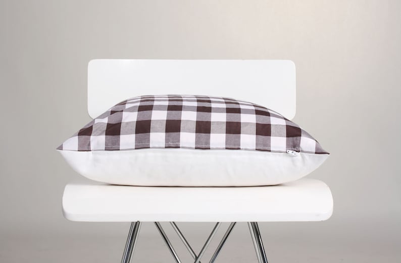 May include: A rectangular pillow with a brown and white checkered pattern on top, resting on a white pillow base. The pillow is placed on a white chair with metal legs, against a neutral background.