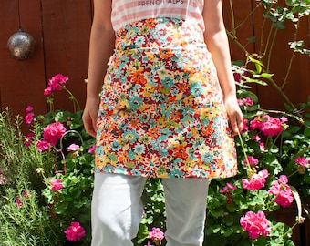 May include: A colorful floral print apron with a red background, featuring orange, yellow, and blue flowers. The apron is worn over light-colored jeans and a striped t-shirt with the text "SAVOIE FRENCH ALPS".