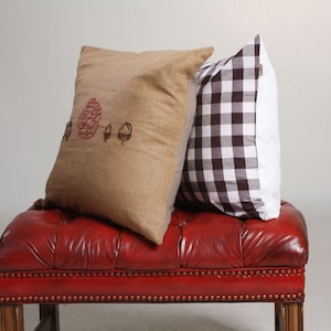 May include: Two decorative throw pillows rest on a red leather bench. One pillow is tan with a red and black acorn design. The other pillow has a brown and white checkered pattern.