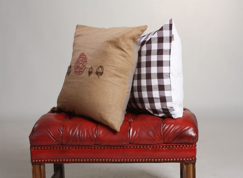 May include: Two decorative throw pillows rest on a red leather bench. One pillow is tan with a red and black acorn design. The other pillow has a brown and white checkered pattern.