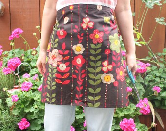 May include: A brown apron with a floral pattern featuring red, green, and cream-colored flowers and leaves. The apron is worn over light blue pants. The background includes pink flowers and a wooden fence.